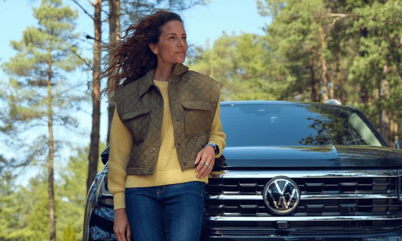 A woman leaning on the hood of a Tourmaline Blue Metallic 2022 Volkswagen Atlas in a forest.