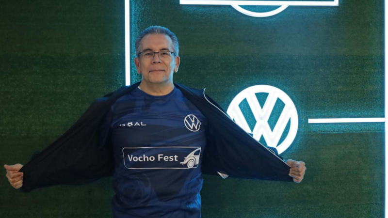 Hombre muestra playera de color azul edición Goal frente a logo de Volkswagen.