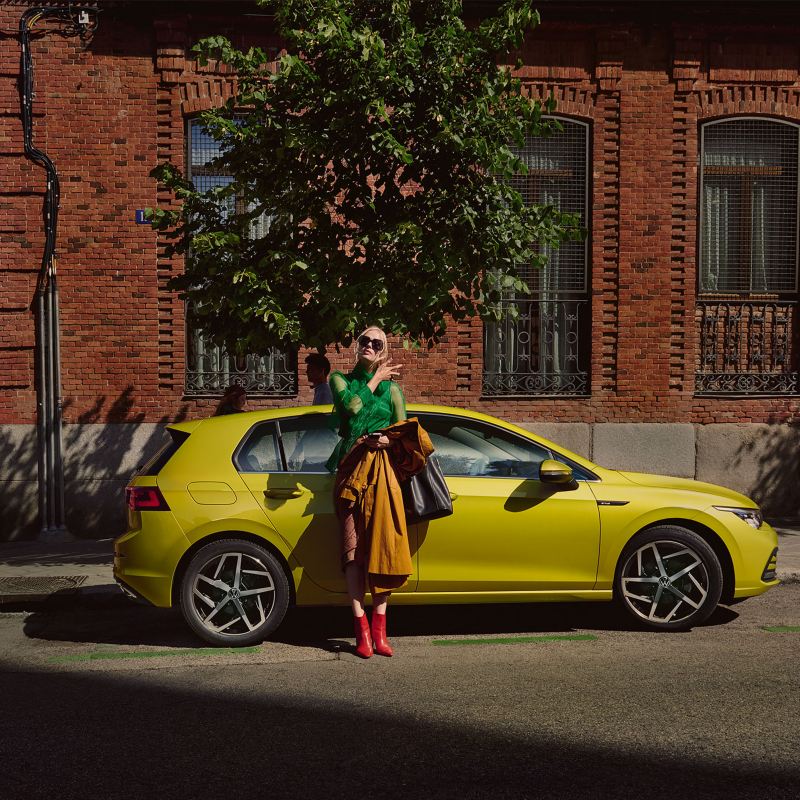 Nouvelle Golf eHybrid jaune devant un arbre et une maison avec une femme appuyée sur la voiture
