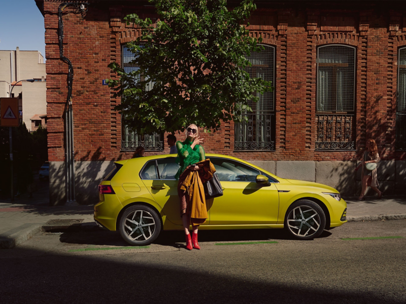 Nouvelle Golf eHybrid jaune devant un arbre et une maison avec une femme appuyée sur la voiture