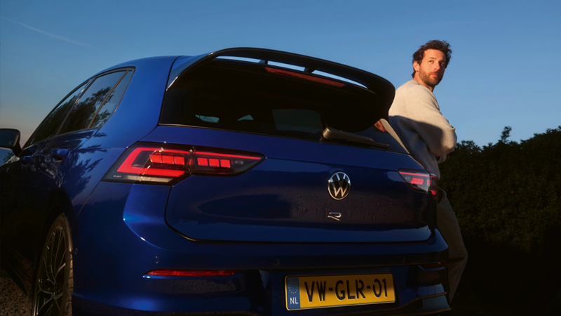 Achteraanzicht van een blauwe Volkswagen Golf R, een man leunt tegen de auto