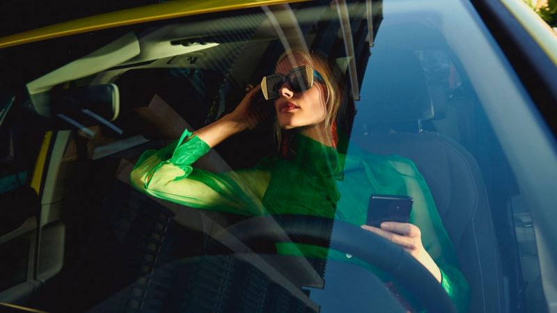 Une femme sur la place conducteur d'une Golf, se regarde dans le rétroviseur, un smartphone à la main.