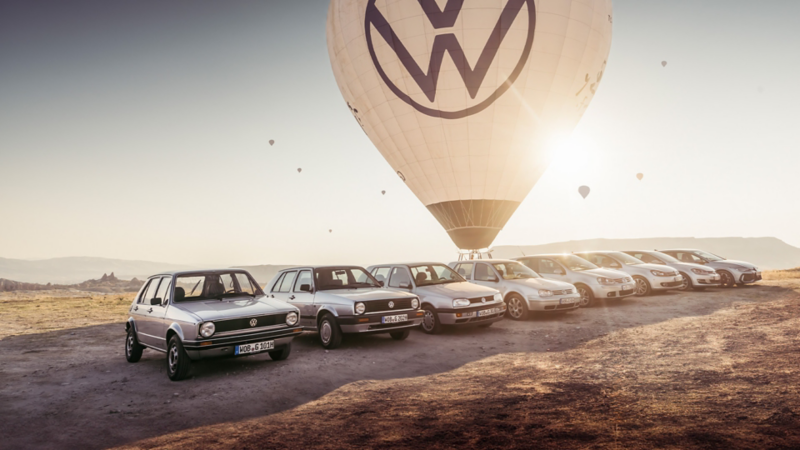 Mehrere Golf Generationen stehen auf einem Platz, während Heissluftballons herum fliegen