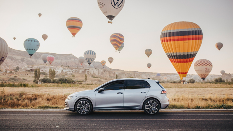 Der neue Golf fährt auf einer Strasse, während Heissluftballons hinter ihm fliegen