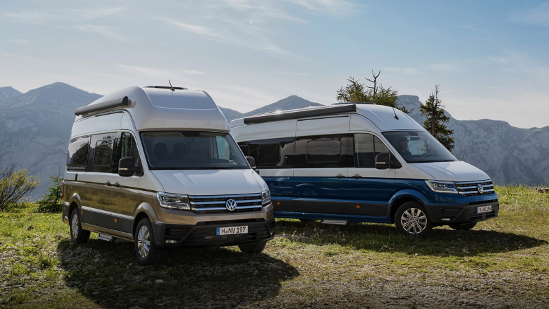  Deux camping-cars Volkswagen Grand California, l'un bicolore beige et l'autre bicolore bleu et blanc, garés dans un paysage montagneux.
