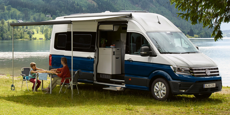 Ein Volkswagen Nutzfahrzeuge Grand California 600 steht am Ufer eines Sees. Die Markise ist ausgefahren. Eine Mutter und ihr Sohn sitzen darunter an einem Campingtisch.