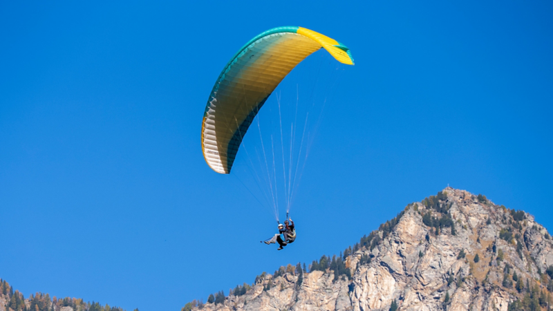 Un parapendio tandem con un vela verde-gialla vola in alto nel cielo azzurro limpido sopra un pendio montuoso boscoso.