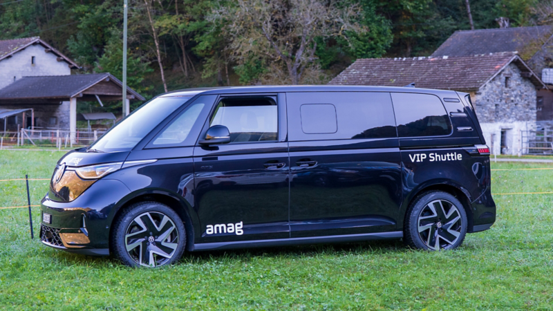 Vista laterale di un ID. Buzz nero con la scritta «VIP Shuttle» e i loghi «amag», parcheggiato su un prato di fronte a vecchie case in pietra.