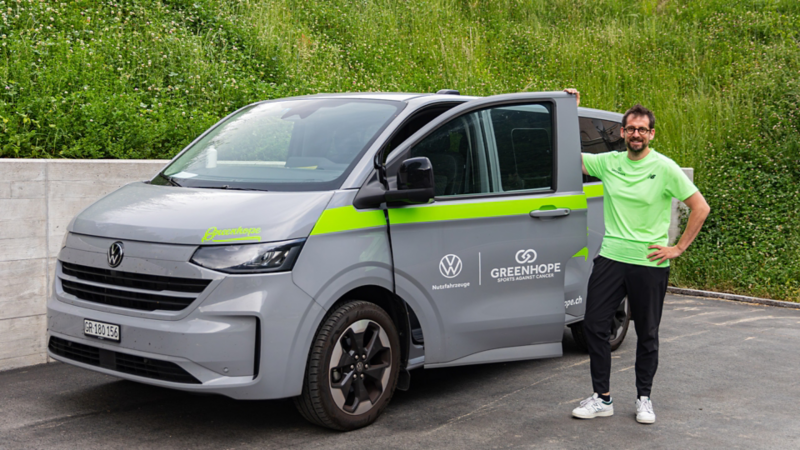 Un uomo con una maglietta verde neon sorride accanto a un veicolo commerciale VW grigio con strisce verdi e loghi di «Veicoli commerciali VW» e «Greenhope».