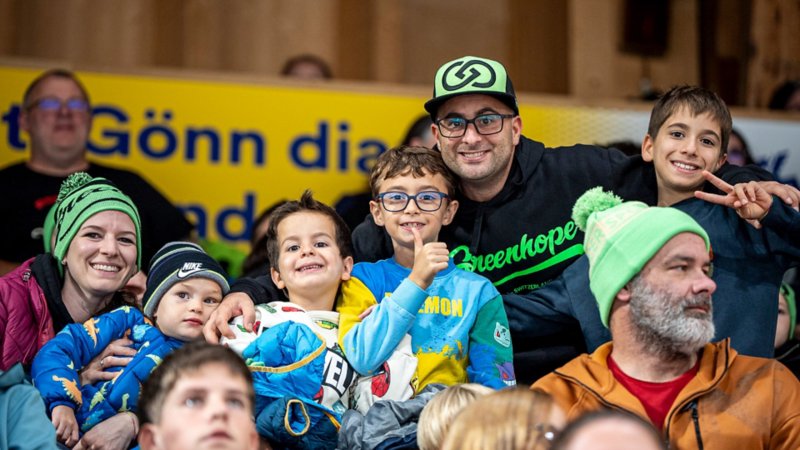 Un gruppo di bambini e adulti che ridono sulle tribune di una pista di pattinaggio sul ghiaccio, molti indossano cappelli verdi o felpe con cappuccio.