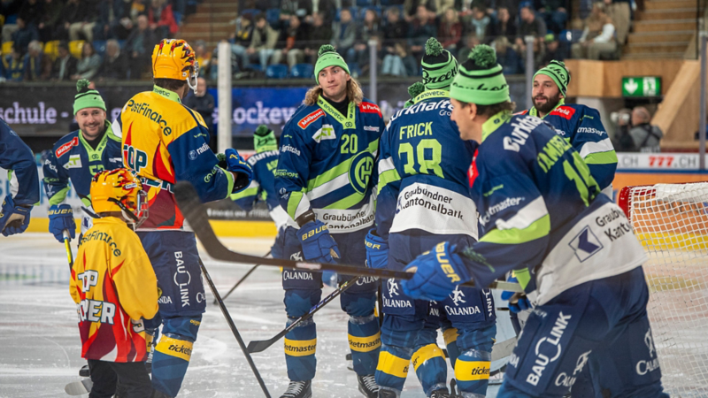 Un gruppo di giocatori di hockey su ghiaccio con magliette blu e cappelli verdi è in piedi sul ghiaccio, alcuni dei quali stanno parlando con un arbitro in una maglietta gialla.