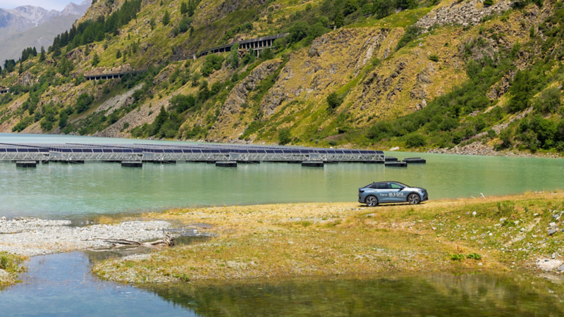 L'ID.5 GTX roule sur un chemin de terre et on peut voir en arrière-plan les panneaux solaires sur le lac des Toules.