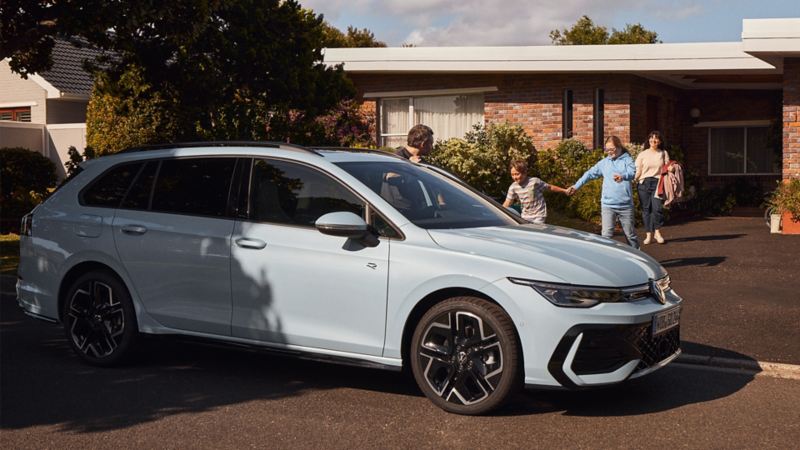 Ein blauer VW parkt vor einem Haus. Eine Familie läuft zu dem Wagen