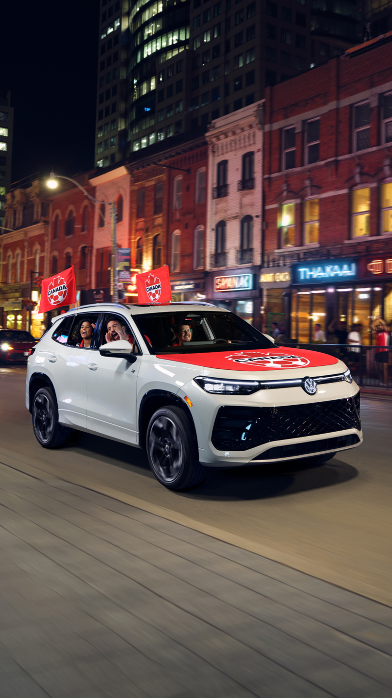 White Volkswagen Tiguan with Canada Soccer flags drives through a lively downtown at night as people cheer from the sidewalk.