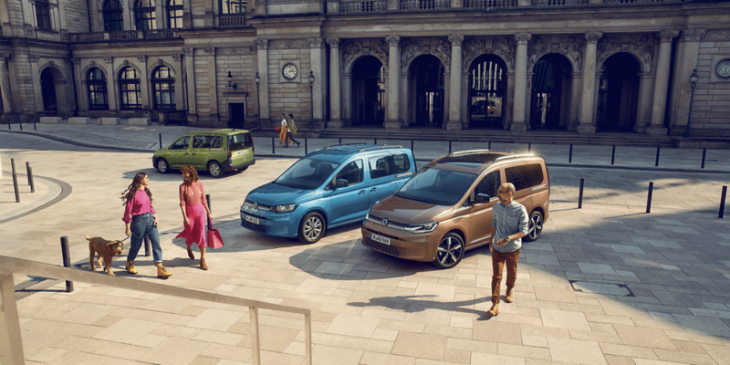 Trois Volkswagen Caddy de différentes couleurs (vert, bleu et bronze) sont garés sur une place pavée. Plusieurs personnes, dont une avec un chien, se trouvent près des véhicules.