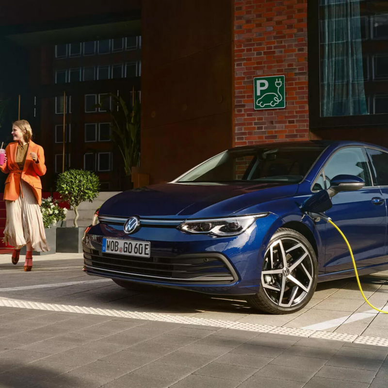 A blue Volkswagen Golf hybrid car is parked in a designated electric vehicle charging spot, plugged into a charging cable, while a woman in an orange blazer walks by holding a drink.