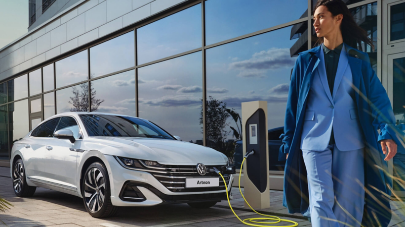 A woman walking away from a white Arteon e-hyrbid that is parked and plugged in to charge.