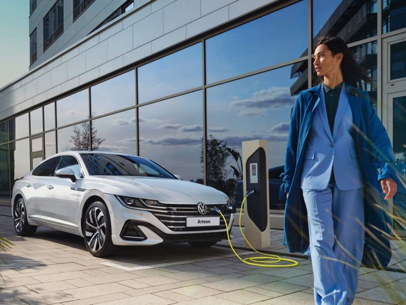 A woman walks past a charging VW Arteon eHybrid in front of a glass façade. 