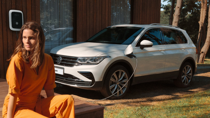 A Volkswagen Tiguan e-Hybrid being charged from a wallbox with a woman sitting closeby