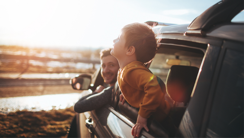 Un père et son enfant, souriants, ont la tête sortie par les fenêtres d’une voiture Volkswagen garée face à une plage ensoleillée. La lumière dorée du soleil illumine la scène.
