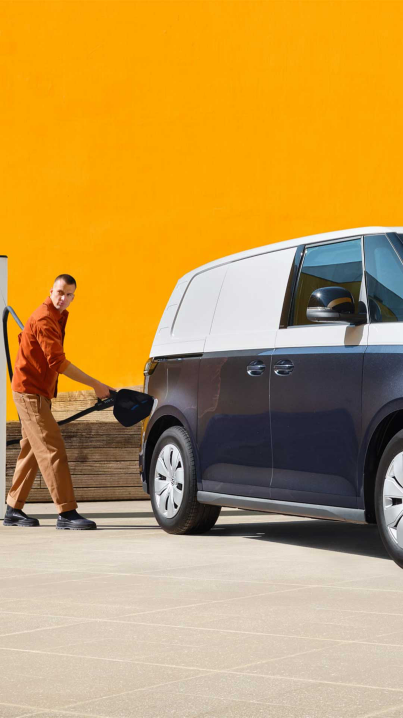 Un homme branche un Volkswagen ID. Buzz Cargo bicolore bleu et blanc à une borne de recharge, tandis qu'une femme se tient à droite du van, tenant une tablette. Le fond est un mur orange vif et un bâtiment moderne.