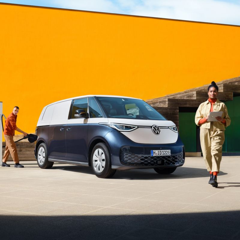 Το VW ID. Buzz Cargo σε μπλε-λευκό, σε έναν σταθμό φόρτισης. Μία γυναίκα στέκεται μπροστά με ένα tablet στα χέρια. Ένας άνδρας συνδέει το καλώδιο φόρτισης.
