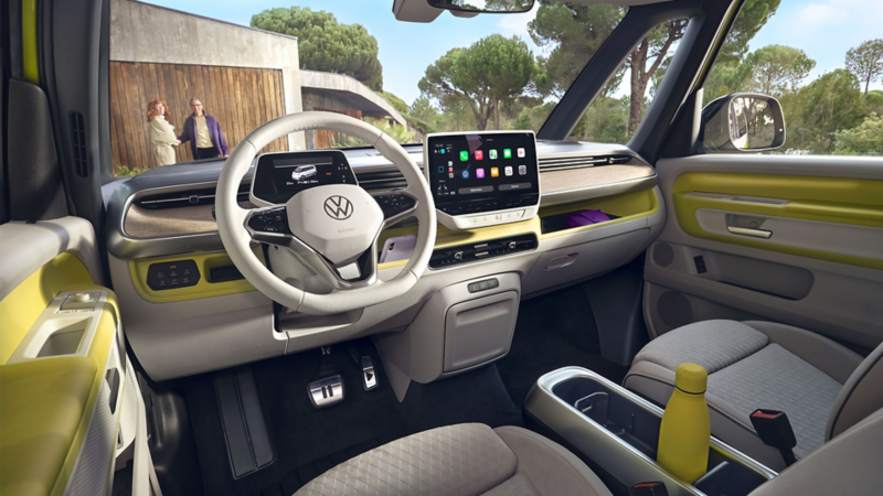 L'intérieur de l'ID. Buzz en jaune clair et gris est présenté ici vu depuis le siège conducteur à travers le pare-brise. Devant l'ID. Buzz, un couple se tient devant une porte de garage.