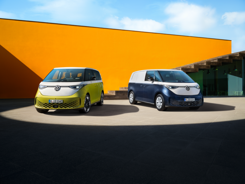 Volkswageny ID. Buzz i ID. Buzz Cargo stoją na placu koło pomarańczowego budynku.