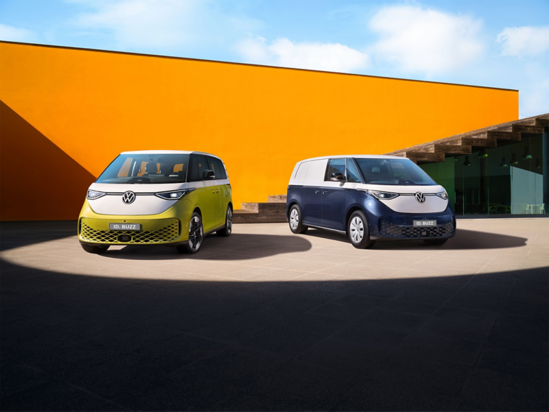 Volkswageny ID. Buzz i ID. Buzz Cargo stoją na placu koło pomarańczowego budynku.