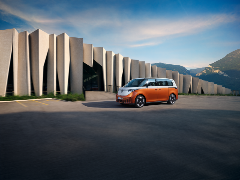 Una furgoneta Volkswagen ID.Buzz bicolor (naranja y blanca) estacionada en una amplia explanada. Al fondo se observa un moderno edificio con pilares verticales y montañas bajo un cielo despejado, combinando diseño y naturaleza.