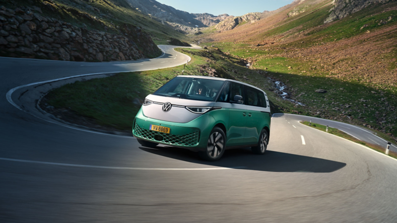  Un Volkswagen ID. Buzz bicolore (vert menthe et blanc) roule sur une route de montagne sinueuse et ensoleillée.
