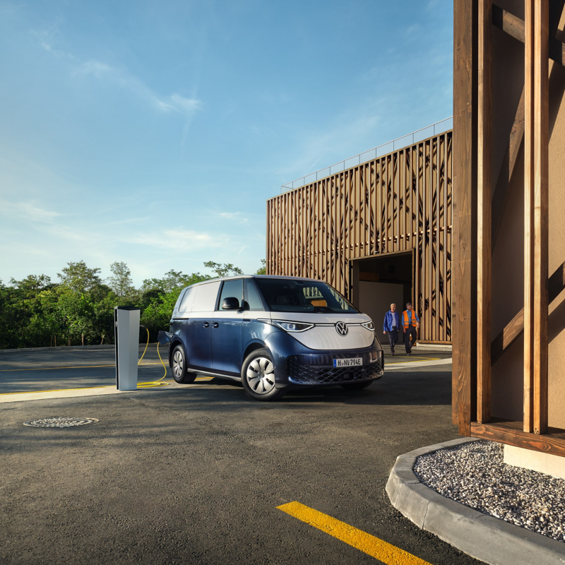 Un Volkswagen ID. Buzz Cargo bicolore bleu foncé et blanc est garé à côté d'une borne de recharge devant un bâtiment moderne avec des panneaux de bois. Deux personnes sont visibles en arrière-plan près de l'entrée du bâtiment.
