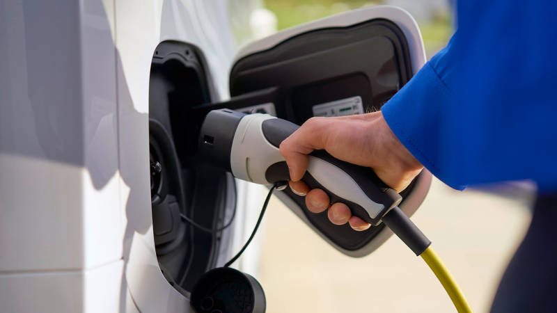 Eine Hand hält einen Ladestecker und steckt ihn in den Ladeanschluss eines weißen Elektrofahrzeugs.
