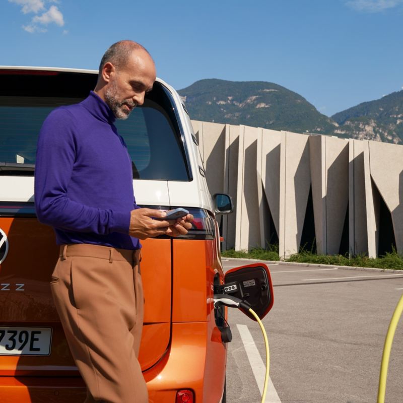 En man laddar elbilen VW ID. Buzz och kollar i sin mobiltelefon