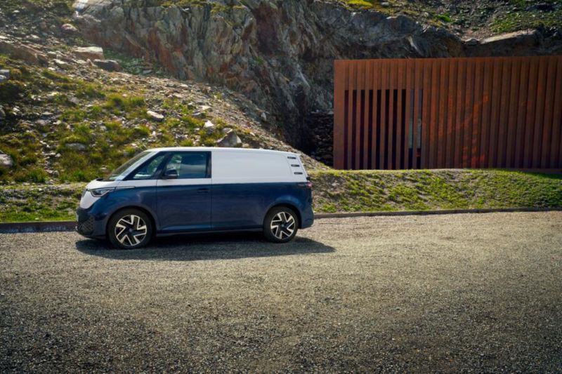 Ein weiß-blauen VW ID. Buzz Cargo steht vor einem Berg auf einem Parkplatz.