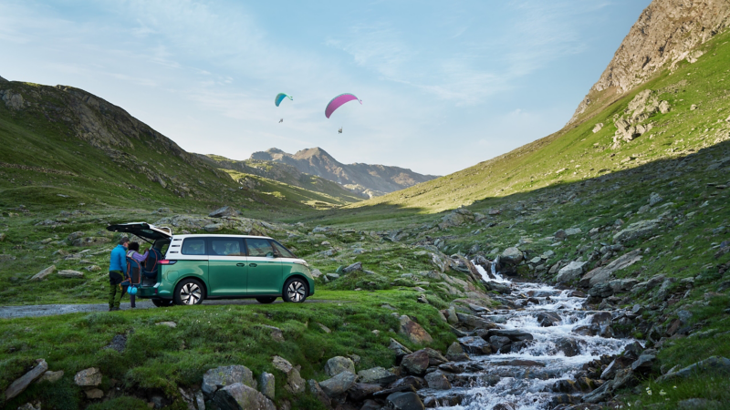 Der VW ID. Buzz parkt an einem Bach mit Bergen im Hintergrund.