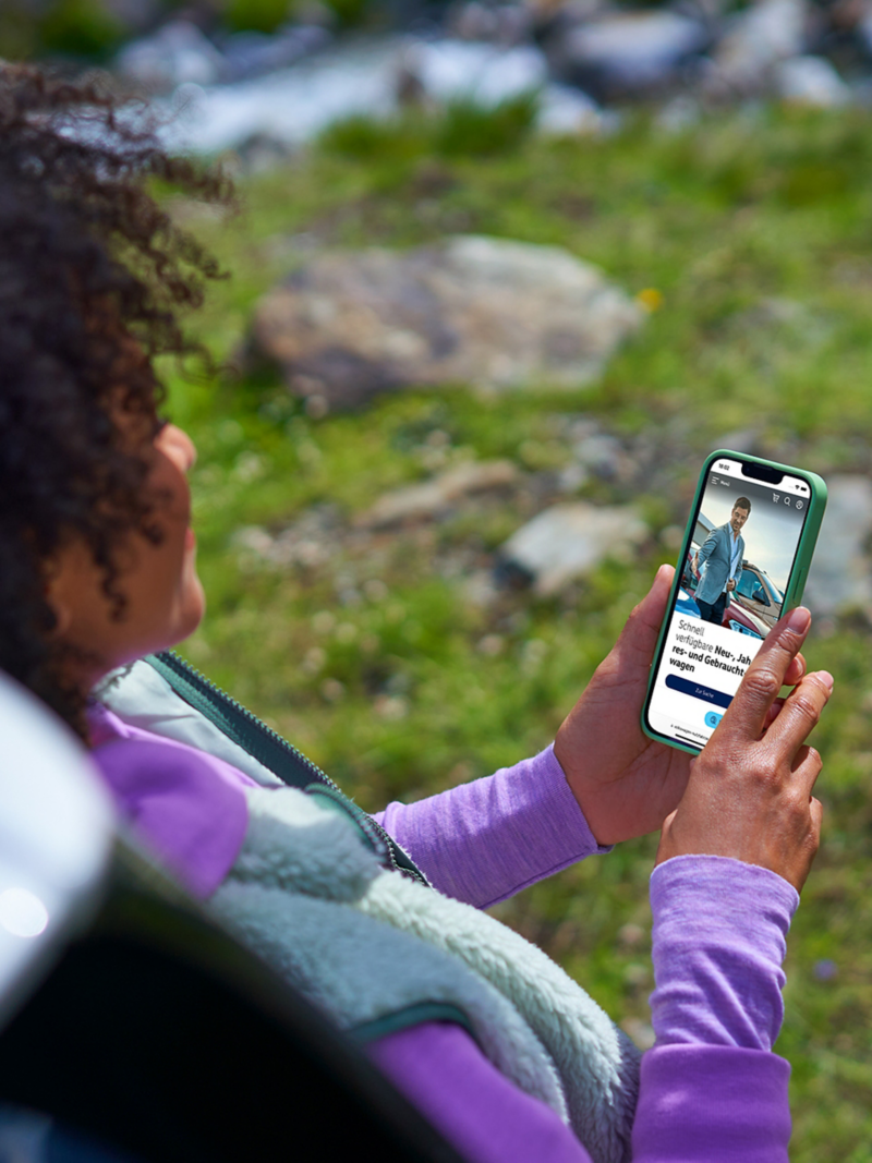 Eine Frau hat ein Smartphone in der Hand, darauf geöffnet ist der VW Online-Shop für Fahrzeuge.