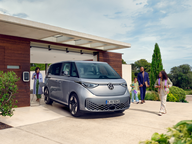 Ein grauer VW ID. Buzz steht auf der Einfahrt eines Hauses an einer Wallbox, eine Familie geht darauf zu.