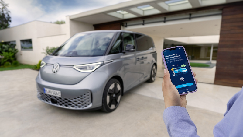 A person holding a smartphone showing the VW Connect app with a vehicle parked outside a house.