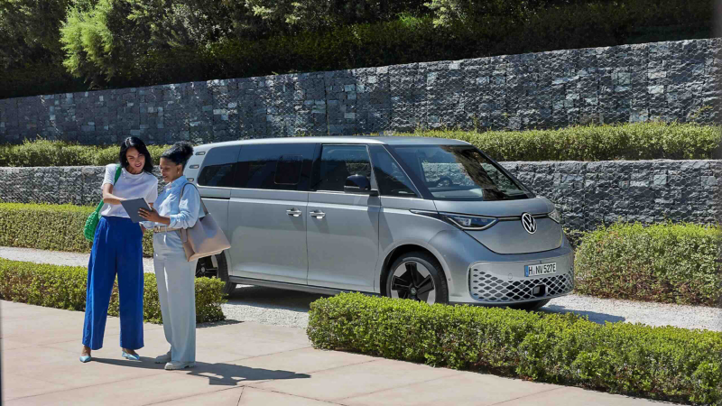 A woman looking at her smartphone. In the background, her Volkswagen is parked in a green car park in a country setting.