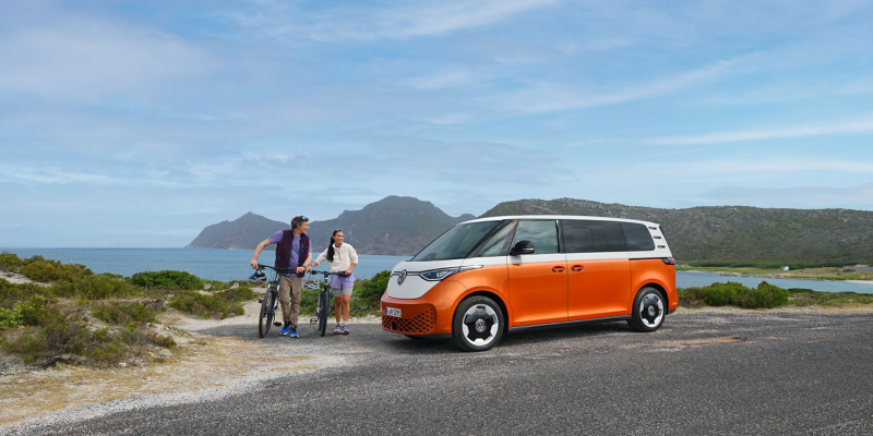 Ein ID. Buzz steht an einem Straßenrand und zwei Personen stehen mit ihren Fahrrädern daneben, im Hintergrund befinden sich Berge und ein See.