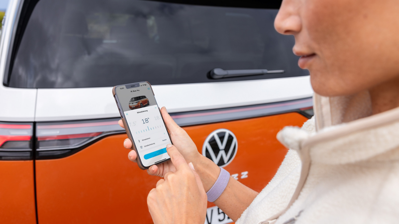 Eine Frau nutzt die Klimatisierungsfunktion einer App auf ihrem Smartphone direkt vor dem Heck eines orangefarbenen VW ID. Buzz.