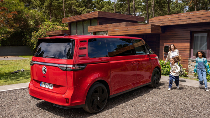 En röd VW ID. Buzz parkerad i på en uppfart med en familj bredvid