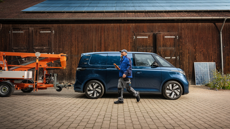 Ein VW ID. Buzz Cargo parkt vor einer hölzernen Scheune mit Solaranlage auf dem Dach. An das Fahrzeug ist ein mobiler Kran gekoppelt. Neben dem Fahrzeug geht ein Handwerker in Richtung des Krans.