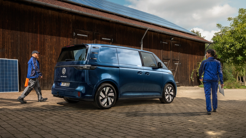Ein VW ID. Buzz Cargo parkt vor einer hölzernen Scheune mit Solaranlage auf dem Dach. Um das Fahrzeug gehen zwei Personen aus dem Handwerk in Arbeitskleidung.