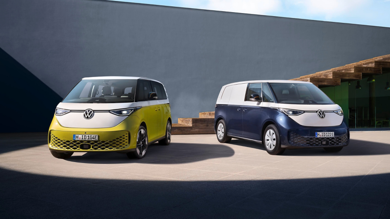 Zwei elektrische Volkswagen ID. Buzz Vans, einer in Gelb und Weiß, der andere in Dunkelblau und Weiß, stehen draußen auf einem gepflasterten Platz.