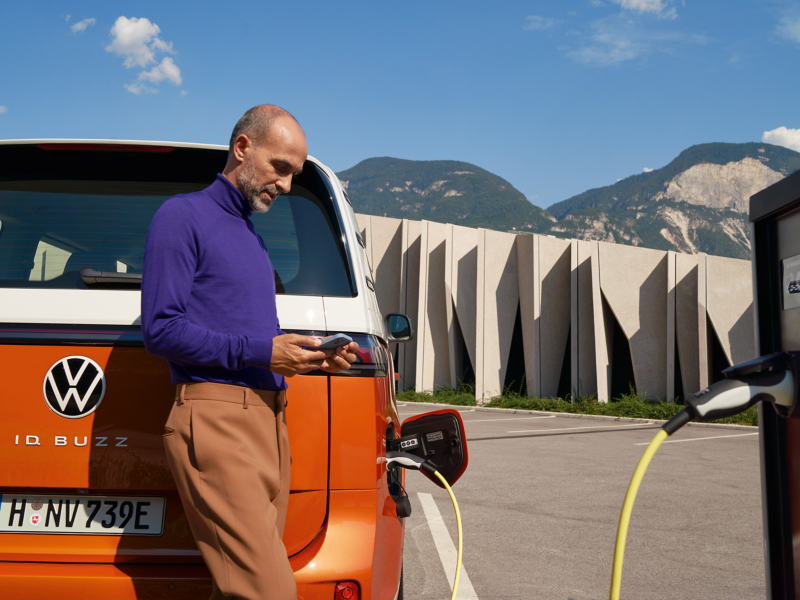 Person standing beside a Volkswagen ID. Buzz while it charges, using a smartphone.