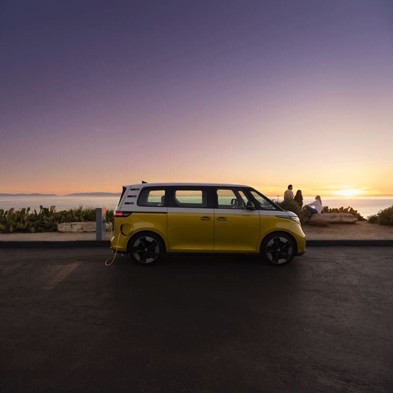 A yellow and white VW ID. Buzz stands to one side on the edge of a beach promenade. In the background, a family with two children looks into the sunset over the sea. 