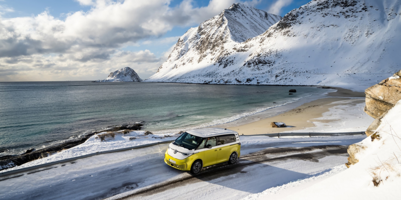 Der VW ID. Buzz fährt durch eine verschneiten Landschaft an einem See, dahinter befindet sich ein Gebirge.