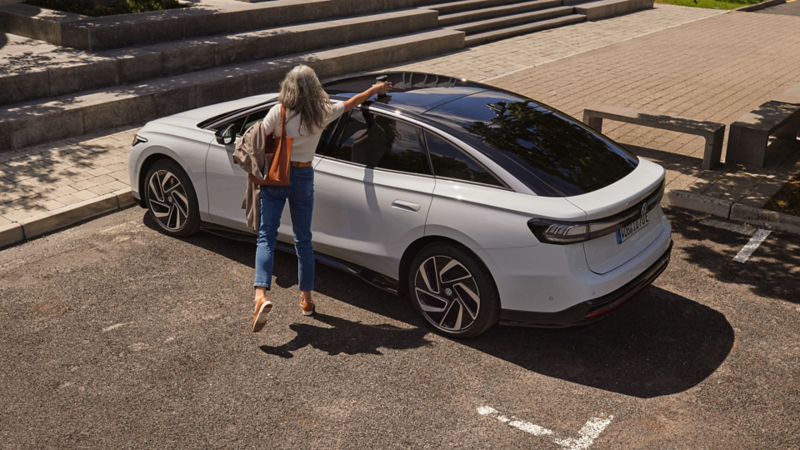 Frau mit weißem Hemd und blauer Jeans steht neben einem weißen VW ID.7. und stellt ihren Kaffeebecher auf das Autodach